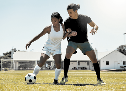 Fotografia de duas pessoas jogando futebol em um campo gramado. À esquerda, uma mulher usa regata, shorts e meias brancas e está com a bola em um dos pés. Logo ao lado dela, está um homem com camiseta preta, shorts cinza e meias pretas.