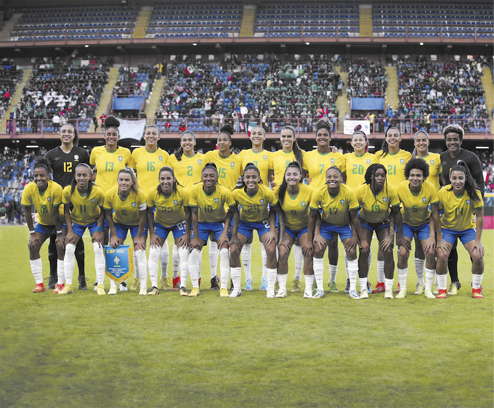 Fotografia da Seleção Brasileira de Futebol Feminino uniformizada, posando para uma foto, em um gramado de um estádio. Elas estão organizadas em duas fileiras: a linha da frente com jogadoras agachadas e a linha de trás com jogadoras em pé. Todas estão usando uniformes do Brasil, com camisas amarelas, calções azuis e meiões brancos. Ao fundo, há arquibancadas com muitas pessoas.