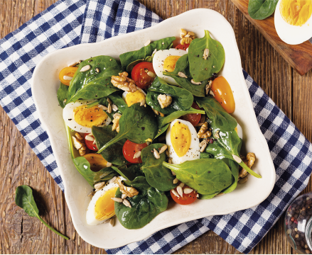 Fotografia de uma salada colorida servida em um prato branco sobre uma mesa com um pano xadrez azul e branco. A salada é composta por folhas verdes, tomates-cereja vermelhos e amarelos cortados ao meio, pedaços de ovo cozido com gema amarela, nozes, e algumas sementes e temperos espalhados por cima.