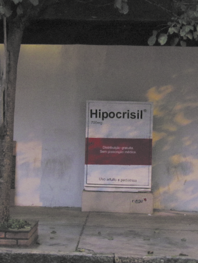 Fotografia de uma instalação artística em uma calçada, composta por uma estrutura que imita a embalagem de um medicamento chamado: Hipocrisil 700 miligramas. Abaixo do nome, há os dizeres: Distribuição gratuita. Sem prescrição médica. Uso adulto e pediátrico. A embalagem é predominantemente branca com uma faixa vermelha no centro. Ela está posicionada em frente a uma parede branca, com uma árvore à esquerda.