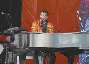 Fotografia de um homem sentado diante de um piano e cantando em um microfone. Ele tem cabelos pretos e bigode, usa uma jaqueta dourada brilhante sobre uma roupa preta.