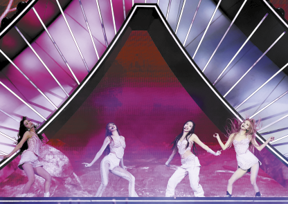 Fotografia de um grupo de quatro mulheres se apresentando em um palco. Elas estão lado a lado, em poses diferentes, usando roupas claras e estilizadas. O palco é grandioso, com uma estrutura iluminada que forma um arco elevado e com o fundo preenchido com cores predominantemente rosa e roxo.