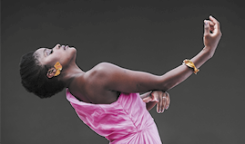 Fotografia de uma mulher durante um movimento artístico. Ela está inclinada para trás, com um braço levemente levantado. Tem cabelos curtos e pretos, usa brincos e pulseira douradas, e usa um vestido rosa.