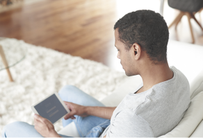 Fotografia de um homem sentado em um sofá branco segurando um tablet, com textos na tela. O homem tem cabelos curtos e pretos, usa camiseta cinza e calças jeans.