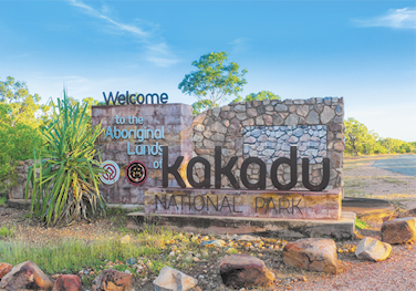 Fotografia de um grande letreiro feito de pedras e concreto próximo de uma estrada. Nele está escrito em inglês: Welcome to the Aboriginal Lands of Kakadu National Park. Ao redor há vegetação com plantas rasteiras e arbustos.