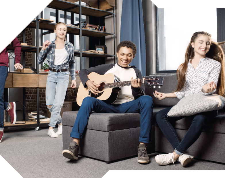 Fotografia de três jovens em uma sala. No centro, um jovem de cabelos curtos cacheados está sentado em um sofá, sorrindo e tocando violão. Ao lado dele, uma jovem de cabelos longos sorri de olhos fechados. Ela está estalando os dedos e com uma almofada no colo. Ao fundo, está uma jovem de camisa xadrez, em pé e sorrindo. Atrás dela, há estantes com vários objetos e livros.