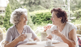 Fotografia de duas mulheres idosas sentadas diante de uma mesa redonda ao ar livre. Ambas estão sorrindo, olhando uma para outro, enquanto seguram xícaras. A mulher à esquerda tem cabelos grisalhos e curtos, e a mulher à direita tem cabelos castanhos avermelhados. Ao fundo há muitas plantas e arbustos.
