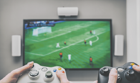 Fotografia de duas pessoas segurando controles de vídeo game de frente para uma televisão que exibe uma partida de futebol.