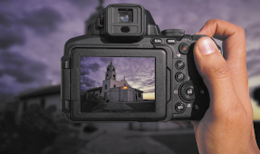 Fotografia de uma mão segurando uma câmera fotográfica e capturando a foto de uma igreja no pôr do sol.