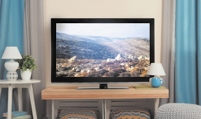 Fotografia de uma sala com uma televisão em cima de uma aparador. Na Tv está sendo exibido a imagem de uma paisagem. Na sala, há cortinas, abajures, pufss e objetos de decoração.