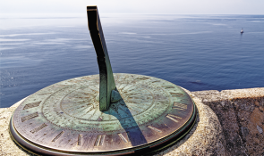 Fotografia de um relógio de sol em uma superfície de pedra, posicionado em um local elevado com vista para o mar. O relógio de sol é circular, feito de metal, e possui números romanos gravados ao longo da borda. No centro, há uma haste triangular projetando uma sombra sobre a superfície do relógio.