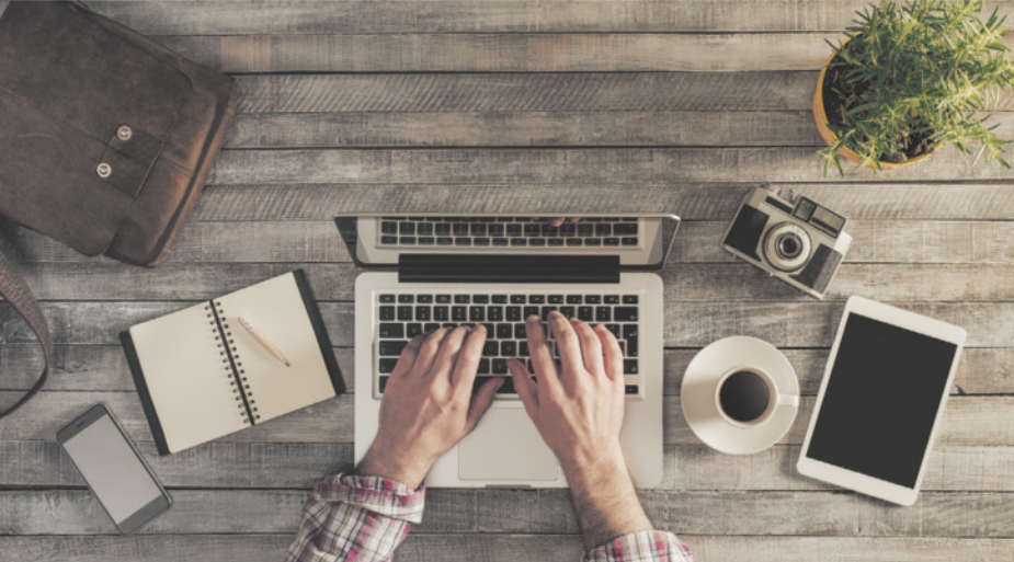 Fotografia das mãos de uma pessoa digitando em um notebook sobre uma mesa. Ao redor há um smartphone, um bloco de notas, uma câmera fotográfica, um tablet e uma xícara de café.