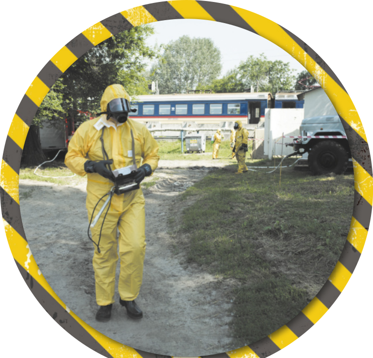 Fotografia de profissionais circulando próximo a uma linha de trem, usando macacão amarelo, luvas, botas e máscara de proteção respiratória. Um deles carrega um medidor de radiação pendurado no pescoço.