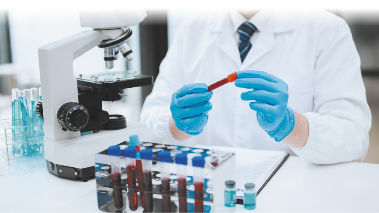 Fotografia de um profissional manuseando tubos de ensaio com sangue em um laboratório. Ele usa luvas e jaleco, e há um microscópio ao lado dos tubos.