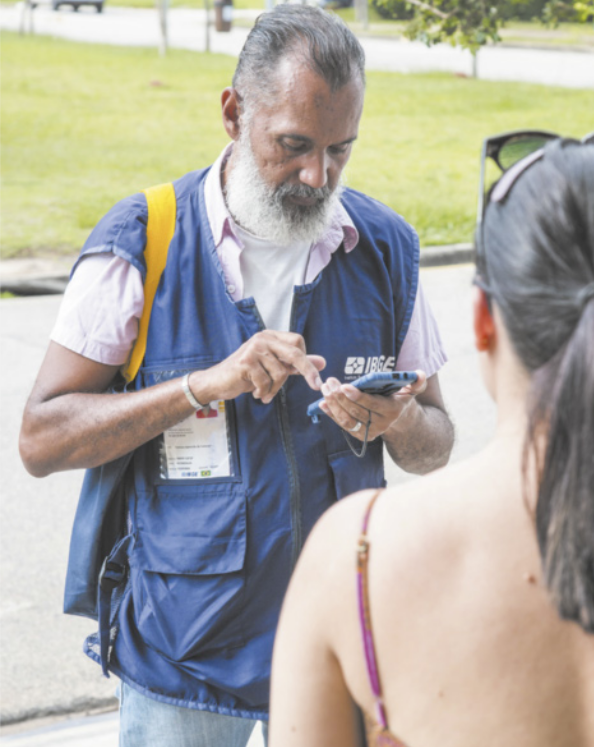 Fotografia de um recenseador utilizando um smartphone para coletar dados de uma moradora. Ele usa um colete com a inscrição I B G E.