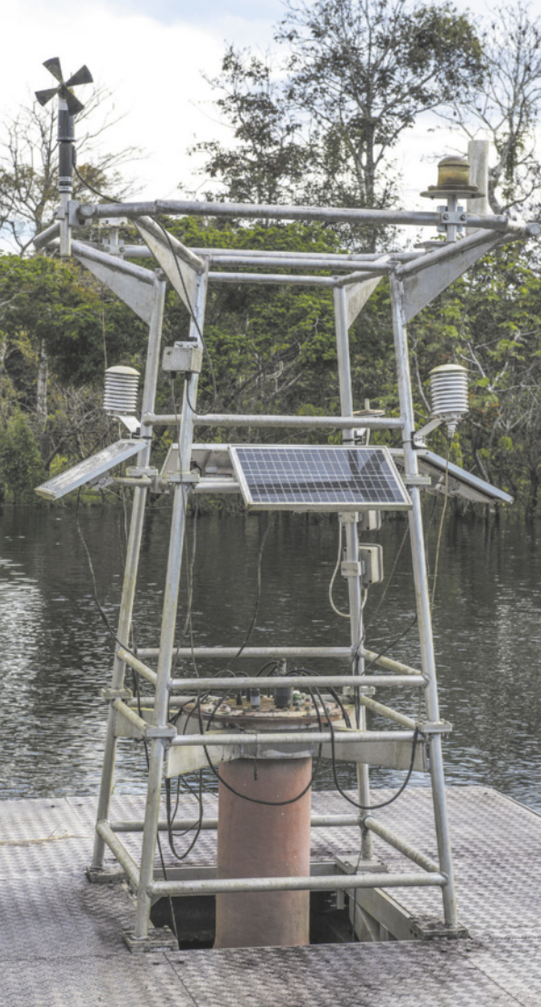 Fotografia de uma estrutura metálica em formato de torre instalada sobre uma plataforma flutuante. Ela é equipada com diversos instrumentos meteorológicos, como anemômetro, pluviômetro, sensores de temperatura e umidade do ar, além de um painel solar. A estrutura está posicionada sobre a água, cercada por vegetação ao fundo.