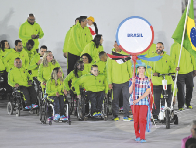 Fotografia de um grupo de atletas em cadeiras de rodas e outros membros da delegação com uniforme verde e preto. Um deles carrega a bandeira do Brasil. À frente, uma jovem com traje típico segura uma placa com o nome Brasil.