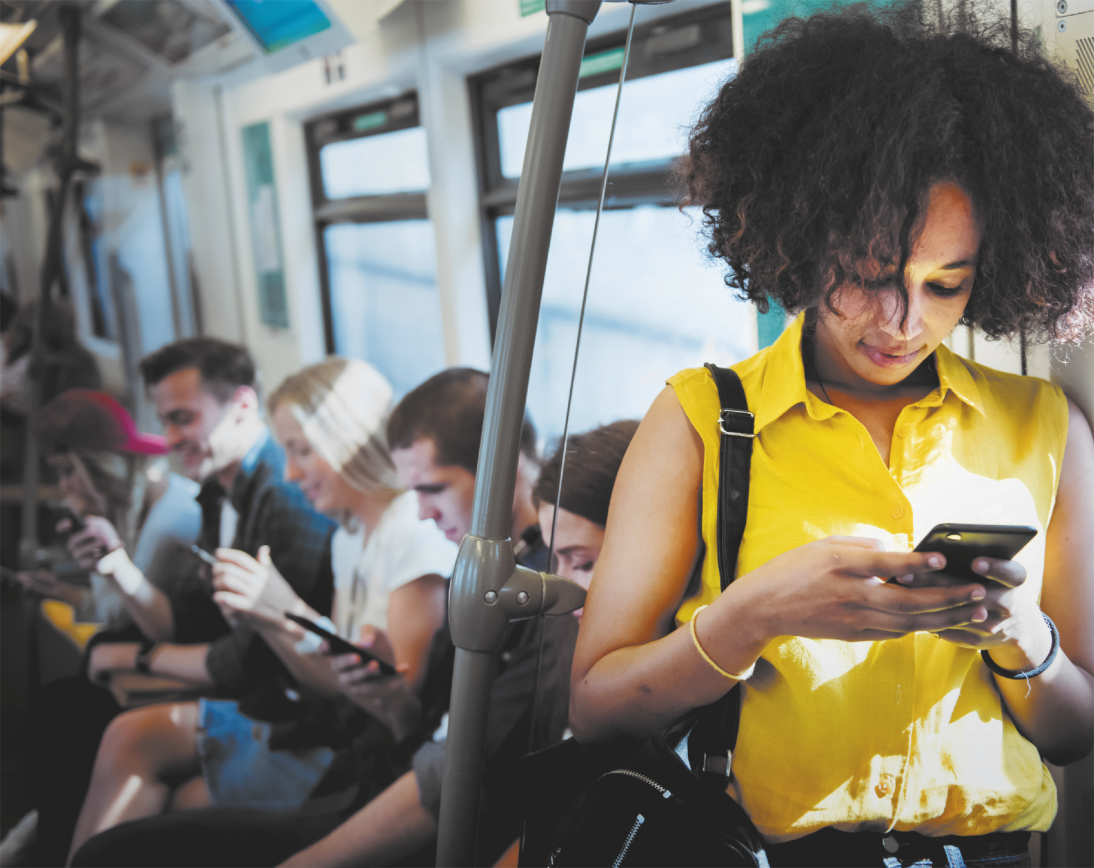 Fotografia de pessoas no metrô, com destaque para uma mulher em pé, enquanto os demais estão sentados. Todos estão focados em seus smartphones.