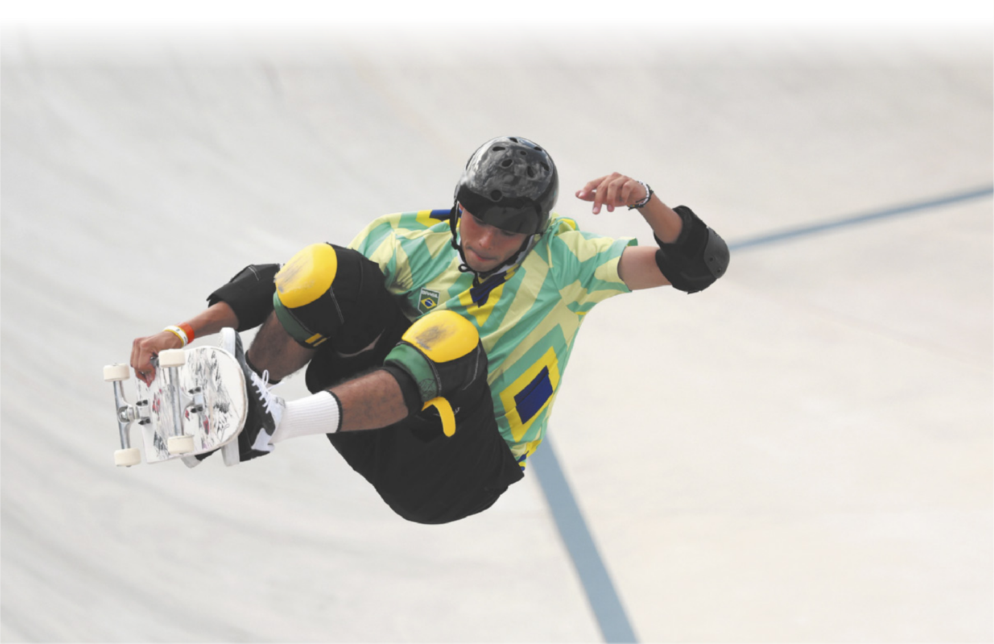 Fotografia de Augusto Akio realizando uma manobra no ar com seu skate. Ele está equipado com capacete, cotoveleiras e joelheiras.