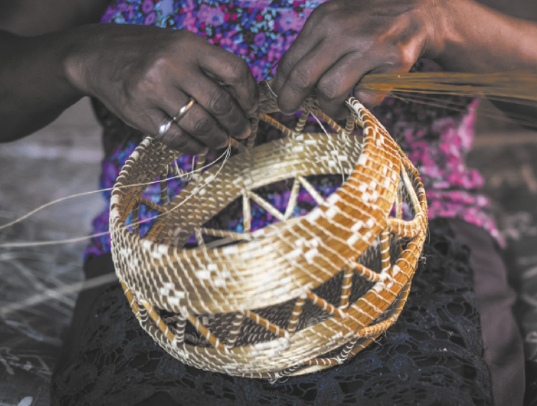 Fotografia das mãos de uma mulher confeccionando um pequeno cesto que apresenta padrões geométricos triangulares.