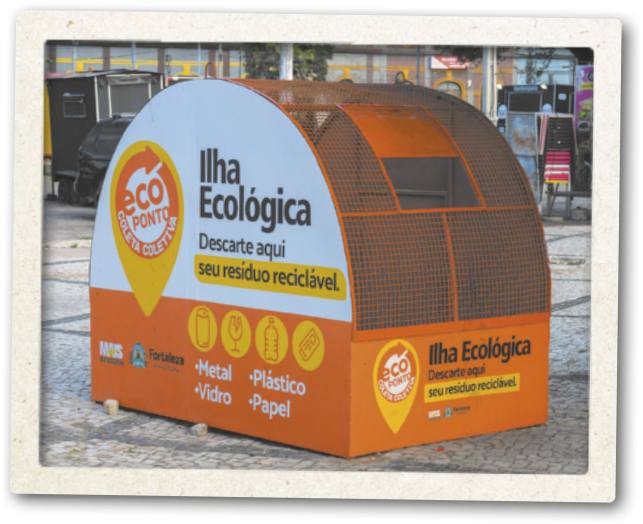 Fotografia de uma estrutura em forma de arco, com um buraco retangular para o descarte de resíduos. Na parte externa temos as inscrições: Ilha Ecológica. Descarte aqui seu resíduo reciclável. Ecoponto. Coleta Seletiva; além de ícones indicando os materiais aceitos: metal, plástico, vidro e papel.