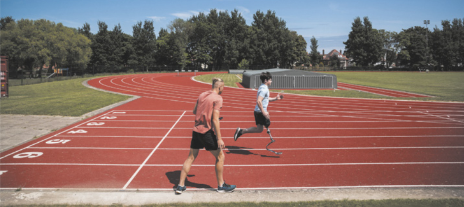 Fotografia de um treinador acompanhando um atleta correndo em uma pista de atletismo, utilizando uma prótese em formato de letra C.