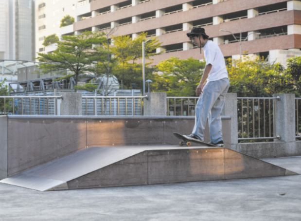 Fotografia de um jovem andando de skate sobre uma estrutura composta por uma plataforma plana e duas rampas em forma de triângulo retângulo, uma de cada lado.