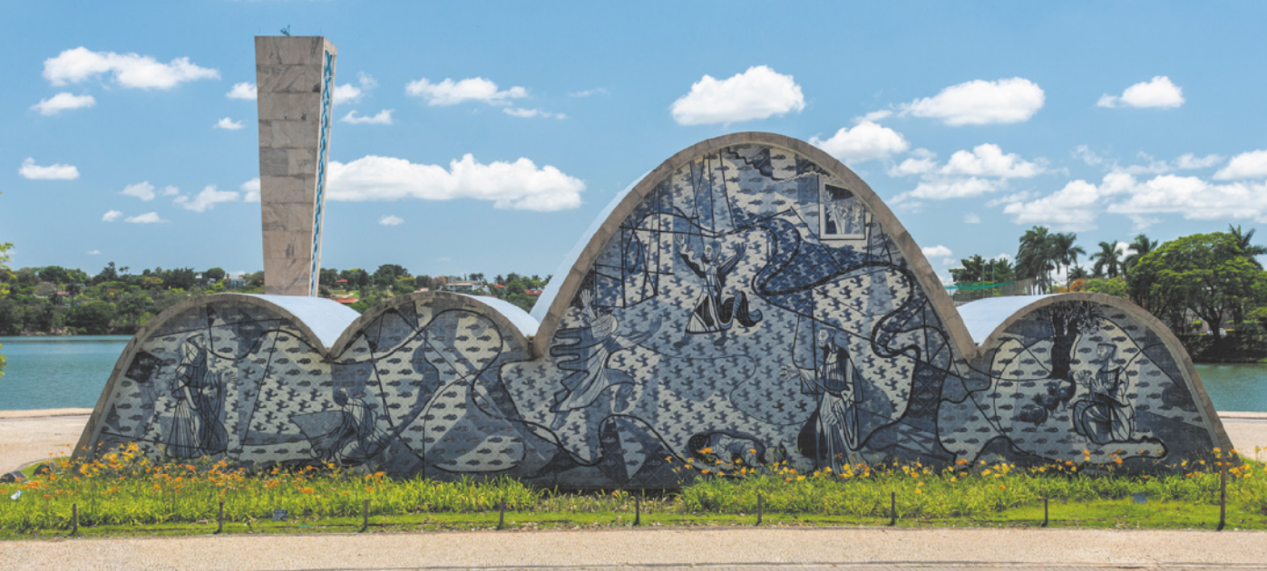 Fotografia de uma estrutura composta por uma torre e quatro abóbadas adjacentes, uma delas mais alta do que as outras, construída próximo a um lago. Sua superfície é revestida por azulejos com figuras humanas em gestos de devoção, andorinhas e peixes.