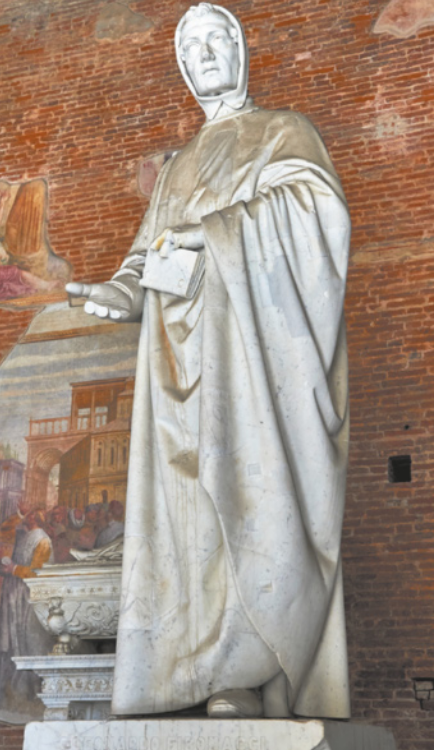 Fotografia de estátua de Fibonacci. Ele usa uma túnica que envolve a cabeça, deixando somente o rosto exposto, e segura um livro enquanto parece dizer algo.