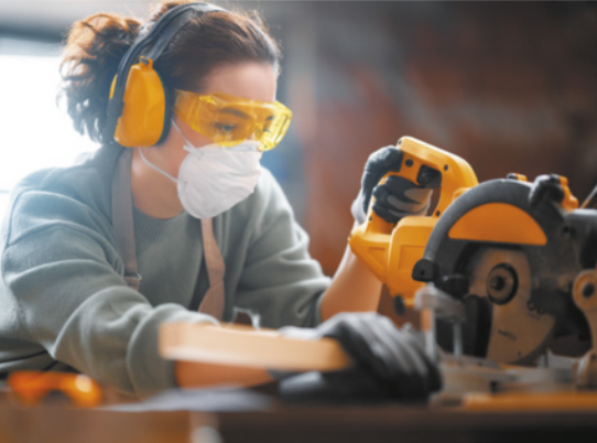 Fotografia de mulher utilizando óculos protetores, luvas e protetores auriculares enquanto manuseia uma serradeira.