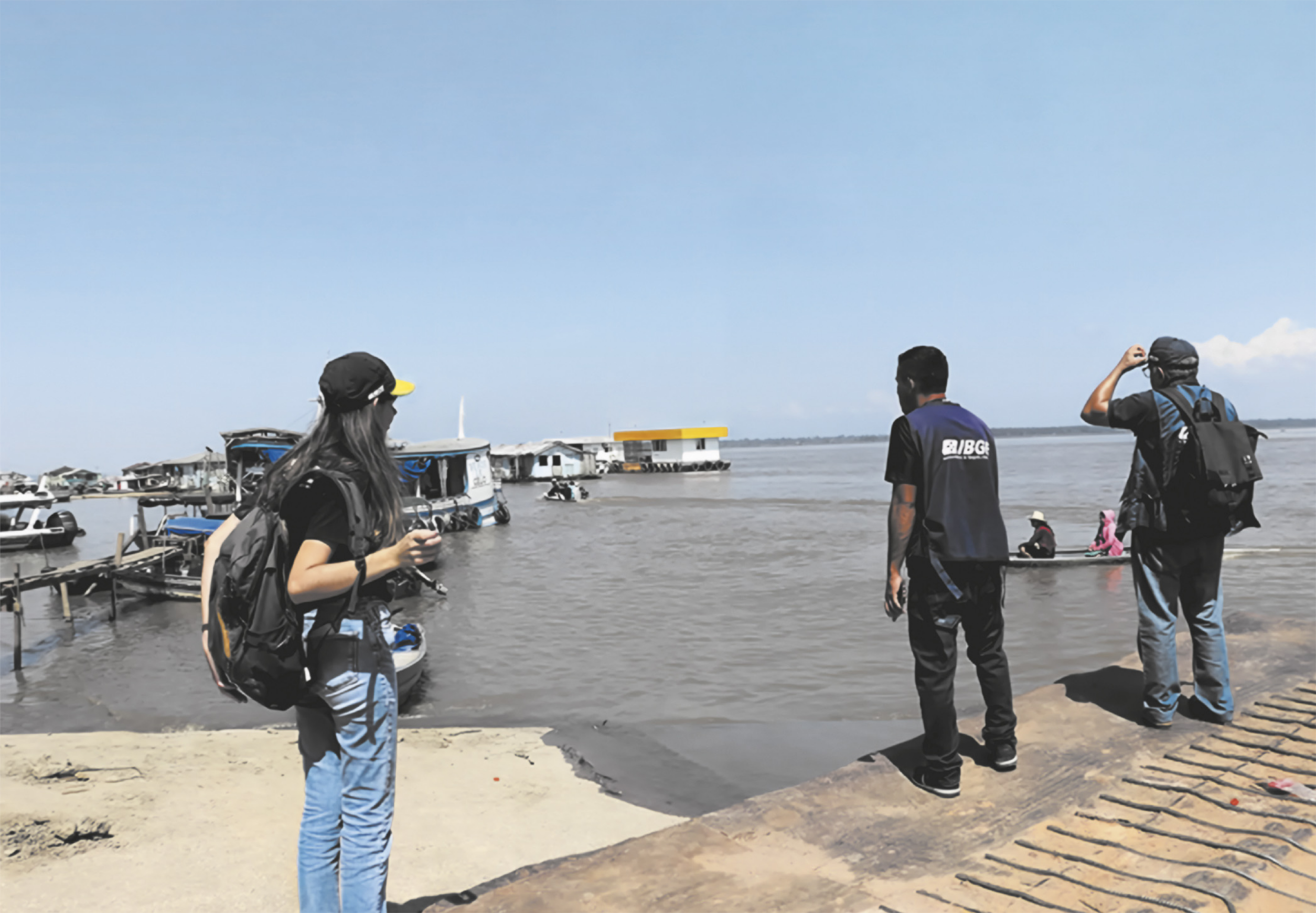 Fotografia de agentes do I B G E em um cais, olhando para a água, na qual há várias embarcações.