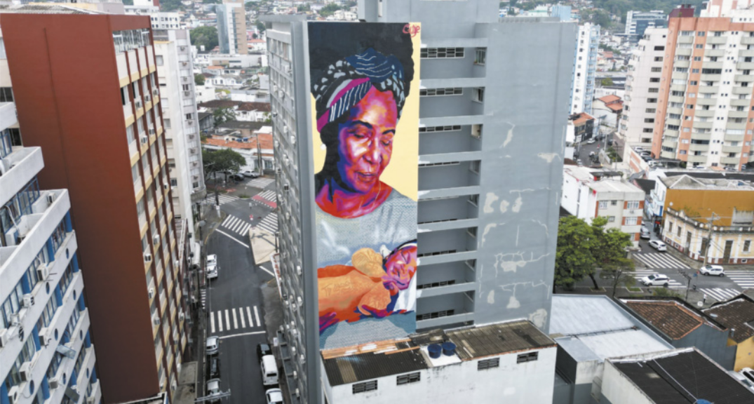 Fotografia com vista de cima de área urbana com muitos prédios. Na parede de um dos prédios há um grande mural representando uma mulher negra segurando um bebê, sorrindo.