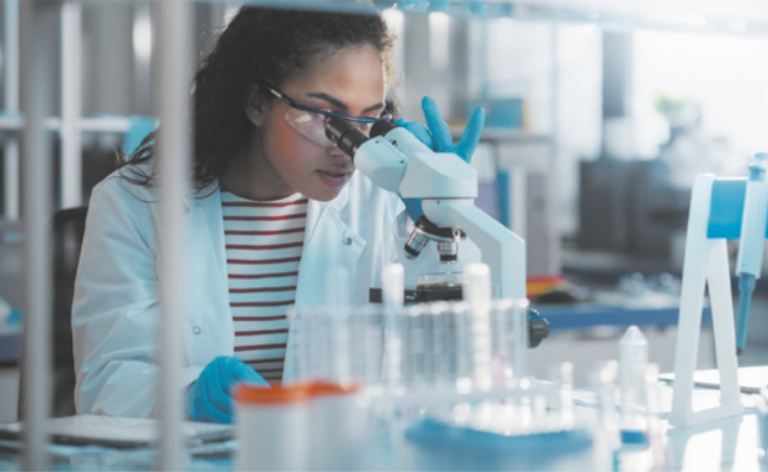 Fotografia de uma mulher em um laboratório. Ela está vestindo um jaleco e óculos protetores enquanto utiliza um microscópio.