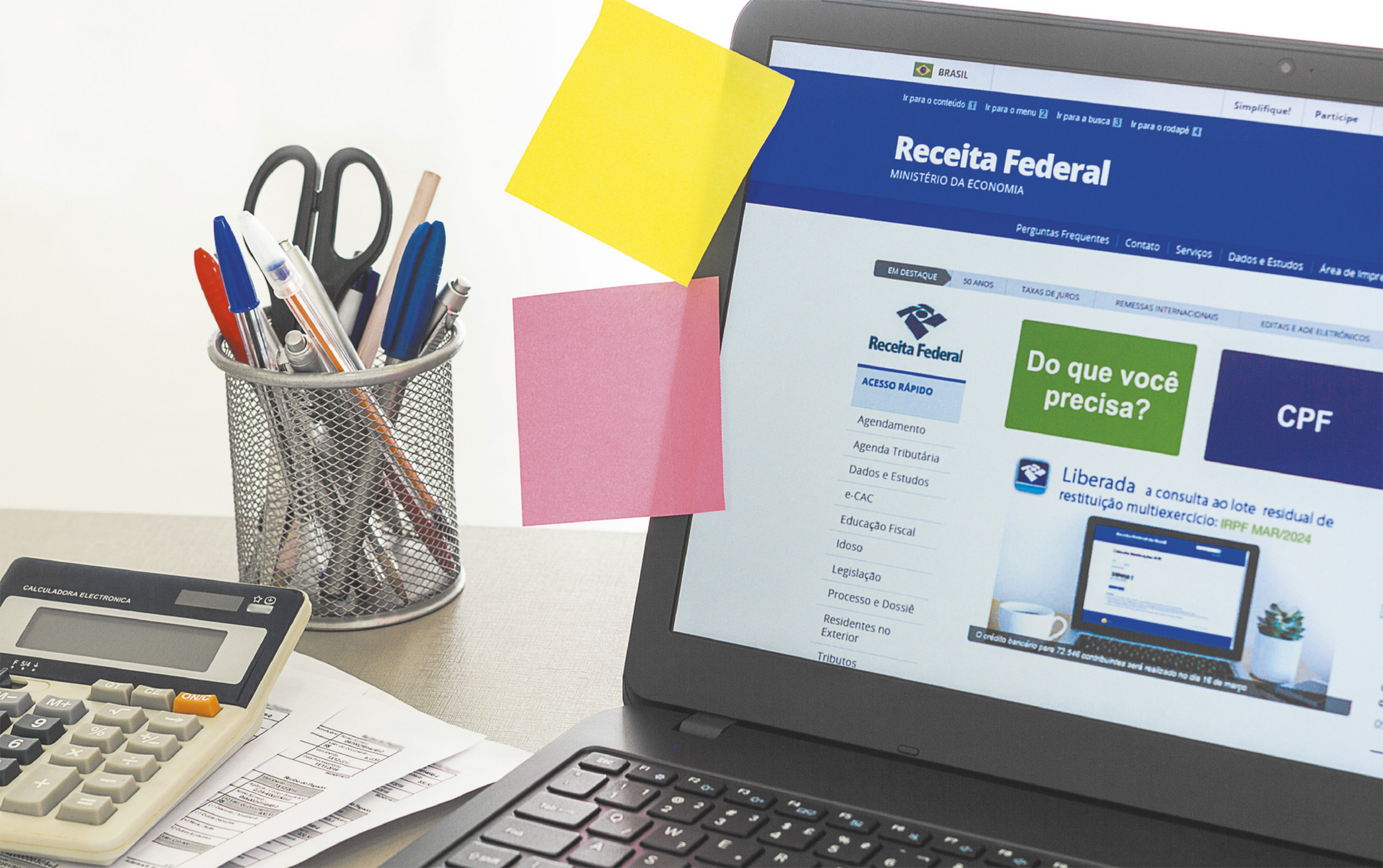 Fotografia de notebook aberto, com o site da receita federal em tela. Ao lado do notebook, uma calculadora, papeis e um porta canetas.