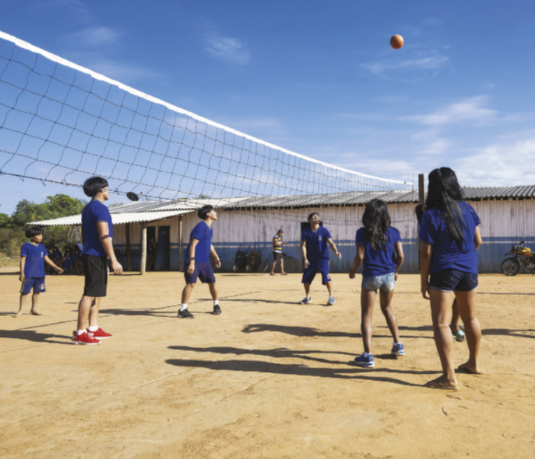 Fotografia de estudantes indígenas jogando vôlei. A quadra tem chão de terra e a escola tem paredes de madeira.