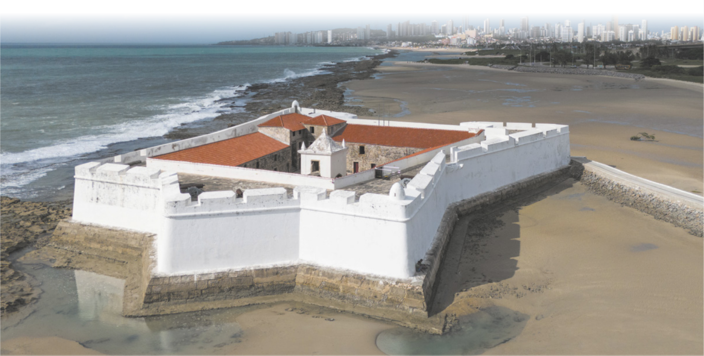 Fotografia com vista de cima de um forte em uma praia urbana. Ao redor dele há areia com algumas poções de água.