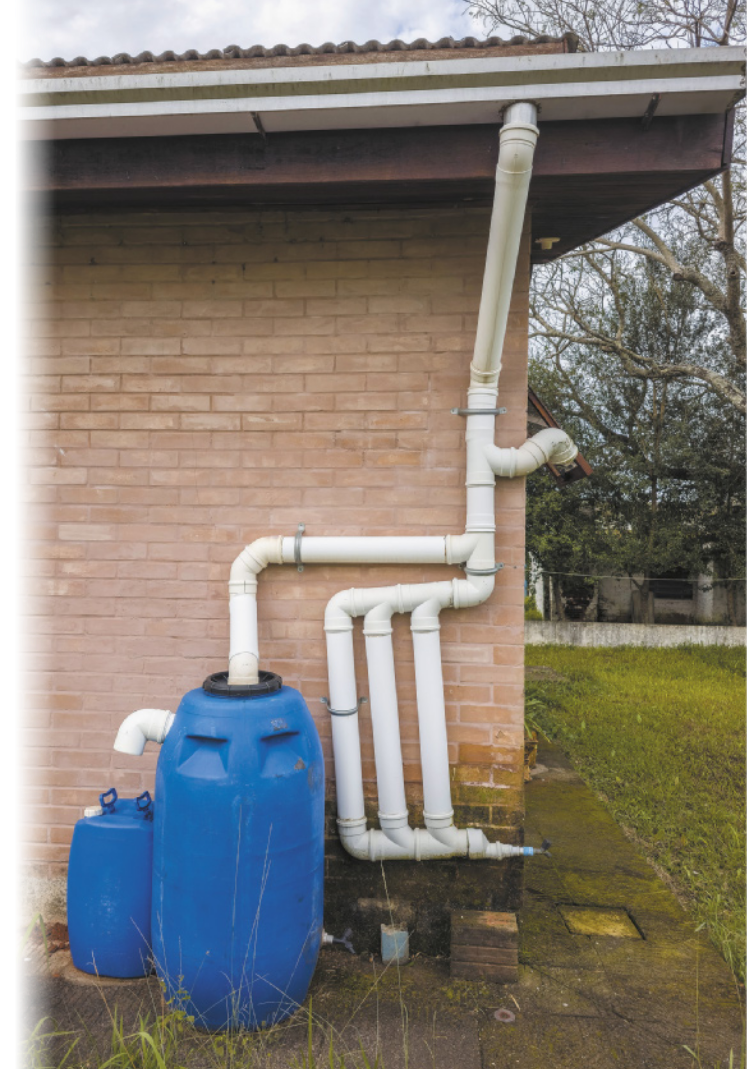 Fotografia de canos conectando o telhado de uma casa a um barril. Um cano principal parte da calha do telhado da casa e ao chegar embaixo há outros canos interligados, alguns indo para a lateral da casa e outro conectado a um barril azul.