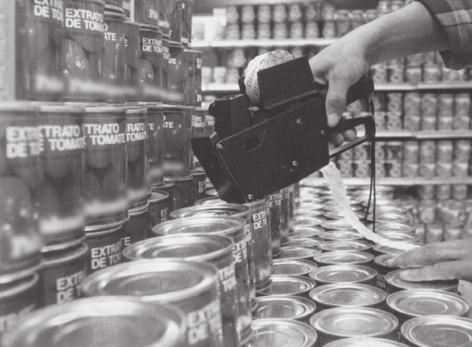 Fotografia em preto e branco de uma etiquetadora e prateleiras cheias de latas.