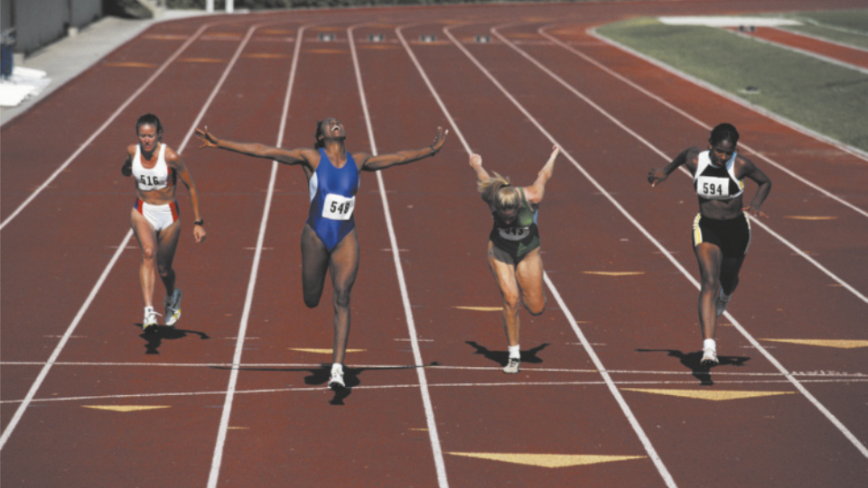 Fotografia de quatro atletas em uma pista de corrida. Uma delas está pouco à frente das demais, tem os braços abertos e olha para cima.