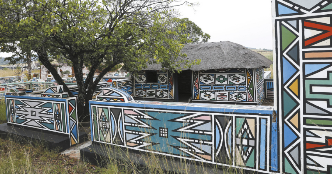 Fotografia de muros baixos pintados com motivos geométricos em cores vibrantes. Há gramado e uma árvore entre eles. Ao fundo há também uma pequena casa cujas paredes são pintadas da mesma forma que os muros.