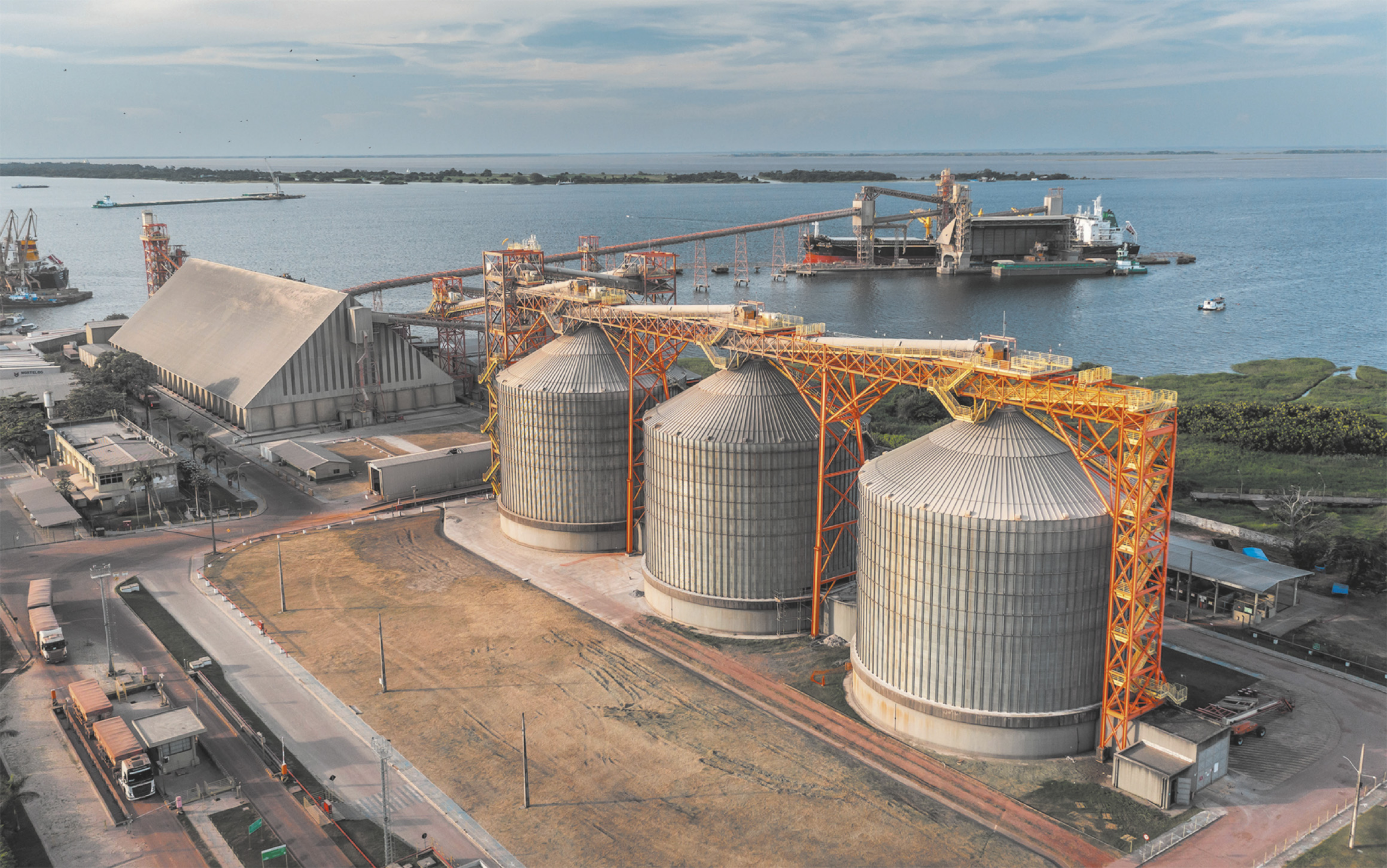 Fotografia de silos em uma fábrica. Os silos possuem formato arredondado com a parte superior em formato cônico. Sobre eles há estruturas de metal de cor laranja.