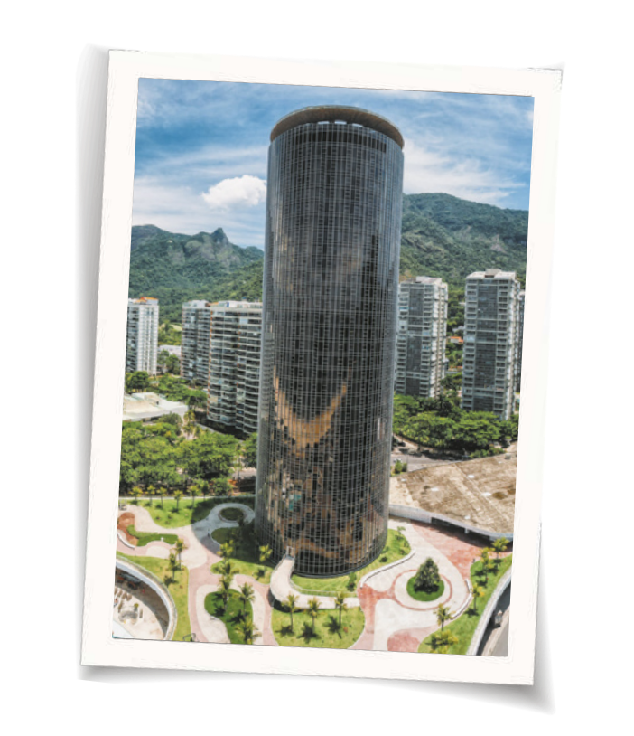 Fotografia de um edifício em formato cilíndrico. Há prédios menores ao redor e, ao fundo, montanhas.