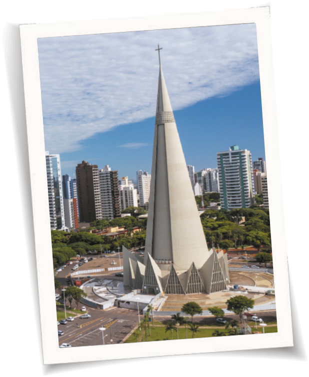 Fotografia de uma catedral em formato cônico. A parte principal possui formato de cone com uma cruz no vértice. Na parte inferior há estruturas triangulares. Está localizada em uma praça circular e há edifícios e árvores ao redor.