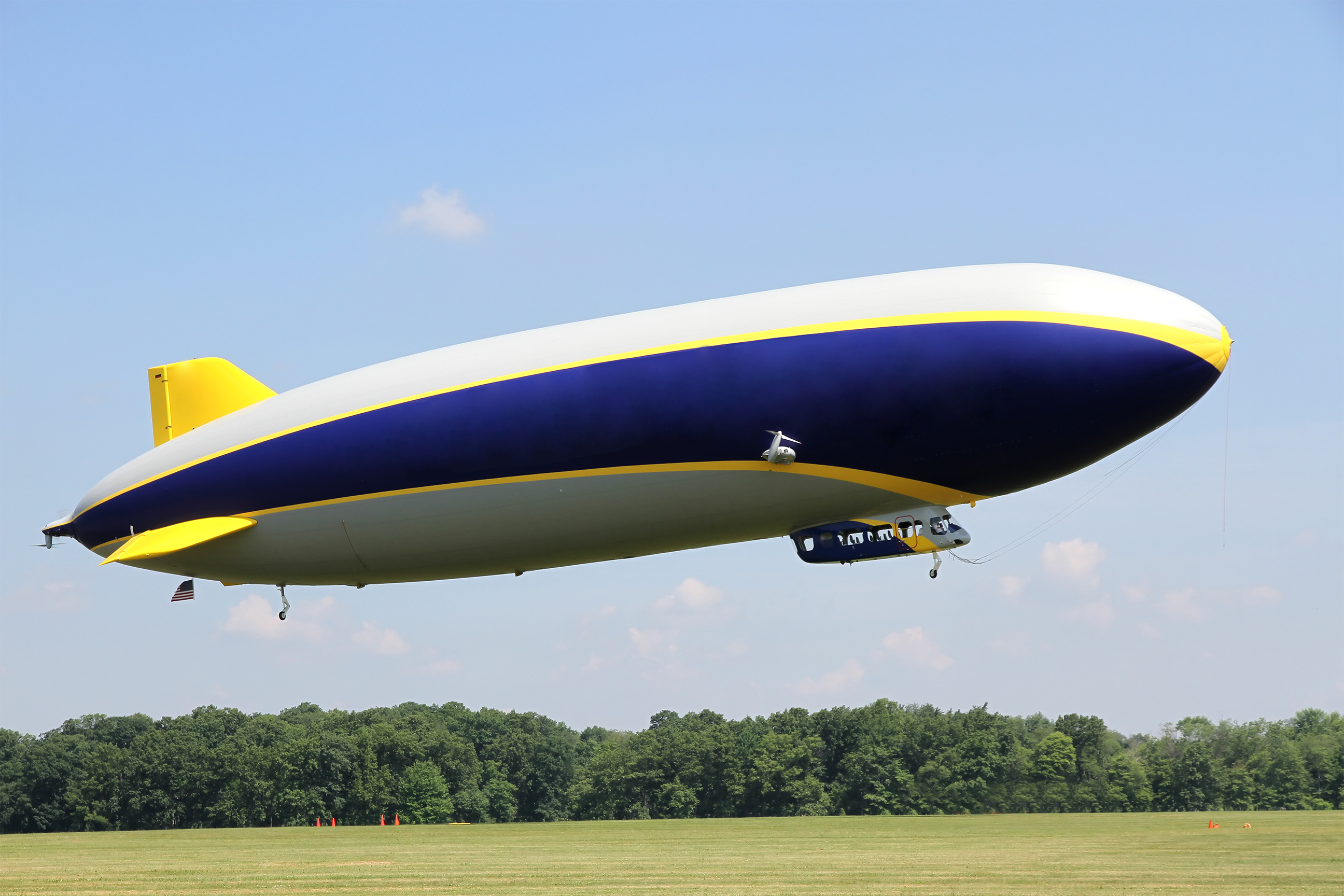 Fotografia de um balão dirigível voando. Ele é uma aeronave nas cores amarelo, azul e branco, com um leme na parte superior traseira e outro na lateral. Na parte inferior traseira, há uma pequena roda; na parte inferior frontal, há um compartimento com janelas e bancos, com mais uma roda embaixo. O dirigível está sobrevoando um gramado com muitas árvores ao fundo. No céu, de cor azul-claro, há poucas nuvens.