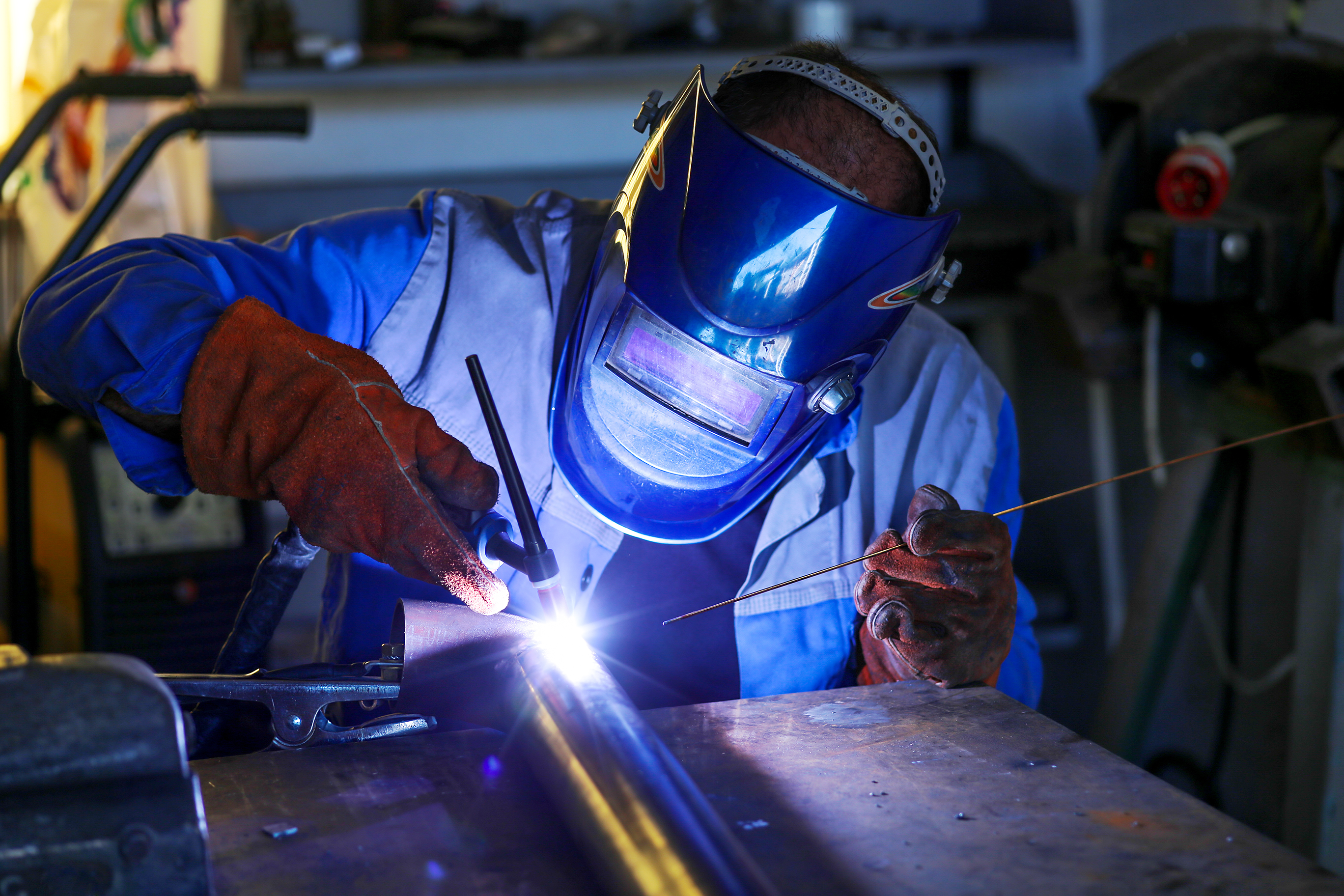 Fotografia de uma pessoa realizando procedimento de soldagem. Ela usa uma máscara com visor cobrindo todo o rosto, um casaco de mangas longas e luvas. Com a mão direita, ela segura um equipamento de soldagem, que está emitindo uma forte luz sobre uma peça que está sobre uma mesa. Na outra mão, ela segura uma vareta de metal.