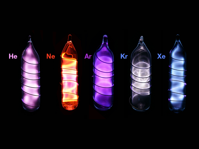 Fotografia de diferentes gases nobres em bulbos de vidro. Eles estão em diferentes cores. Da esquerda para a direita, temos um bulbo com hélio, em fortes tons de roxo; o neônio, em vermelho com partes em amarelo; o argônio, em tons claros de roxo; o kriptônio, em preto e tons de cinza; e o xenônio, em azul.