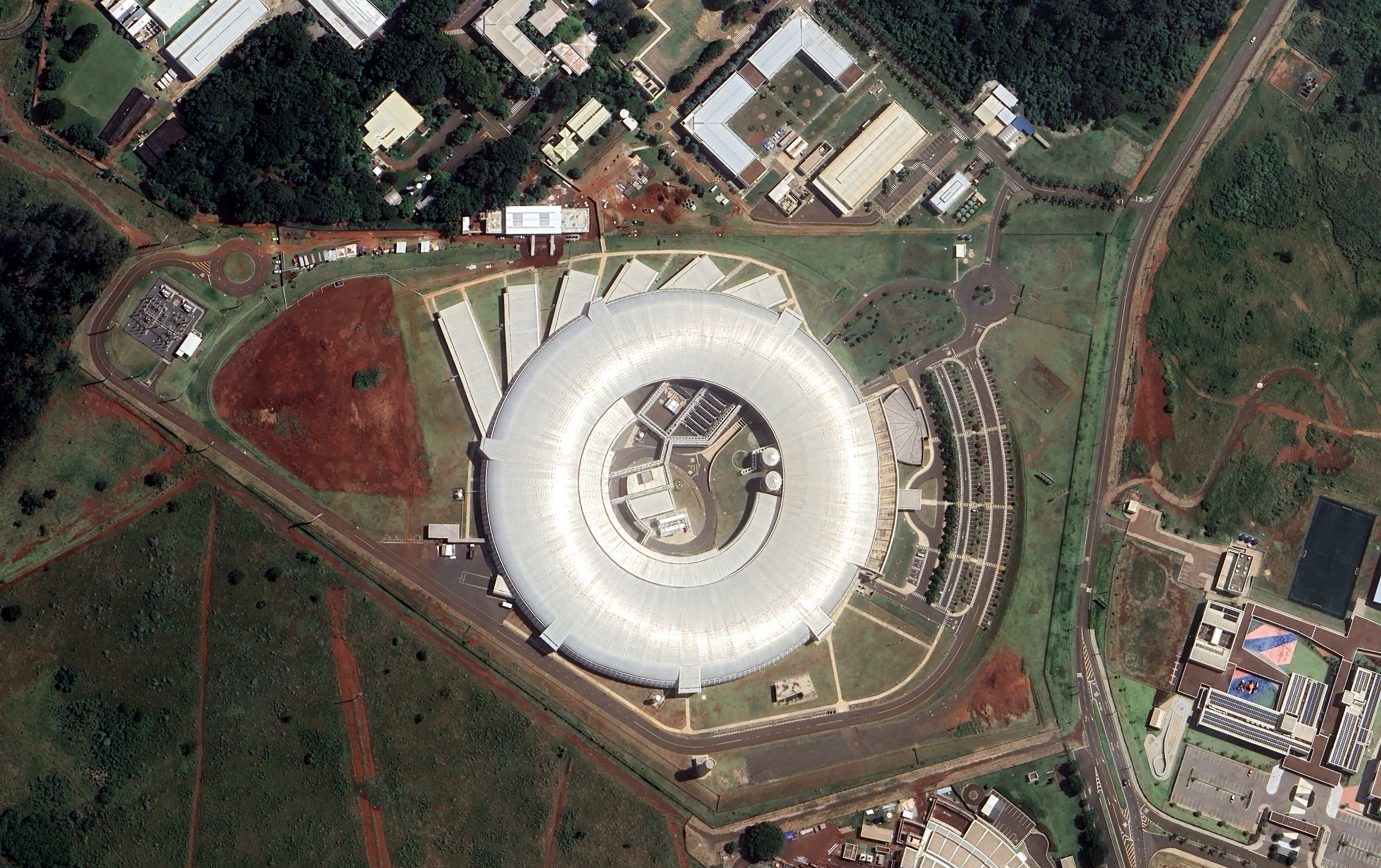 Fotografia da vista aérea de um acelerador de partículas. Ele é uma grande construção em formato circular, sendo semelhante a um estádio de futebol. Ao redor, há muitas áreas verdes, avenidas e outras construções.