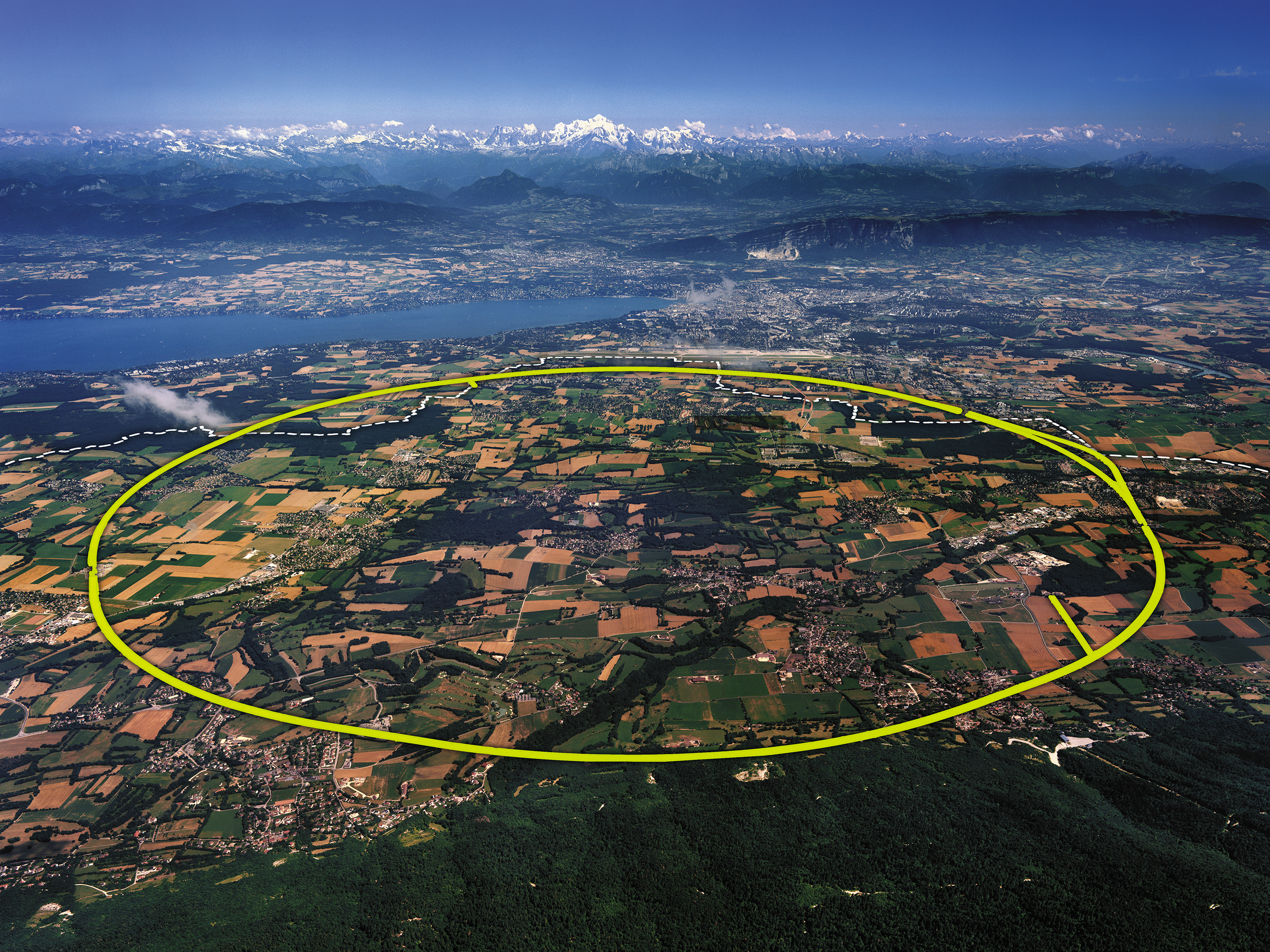 Fotografia da vista aérea de uma paisagem com muitas construções e bastante vegetação. Há uma linha em formato circular desenhada sobre a fotografia abrangendo grande parte da paisagem. Mais ao fundo, há uma área com montanhas.