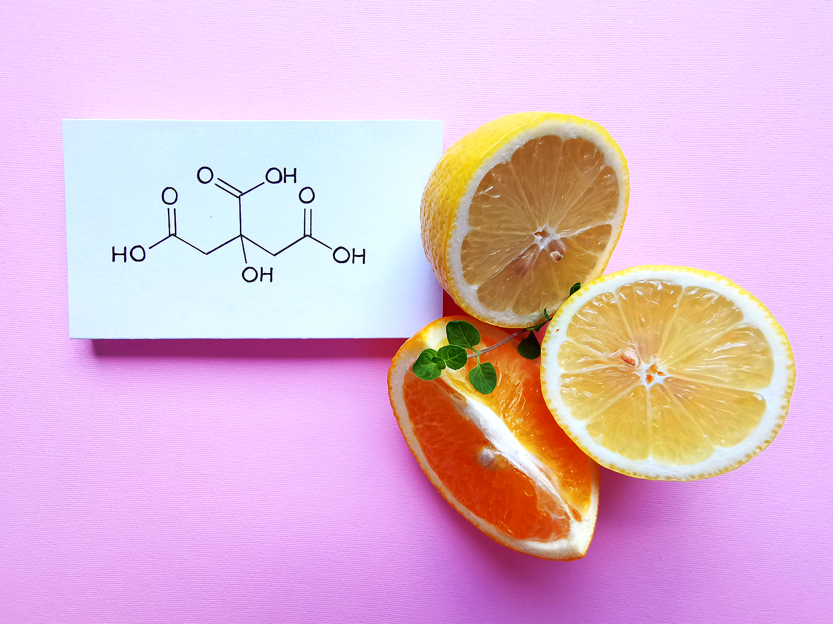 Fotografia de uma laranja cortada, posicionada ao lado de parte de um limão. Ao lado das frutas, há um papel com uma estrutura química que tem as seguintes características visuais: da esquerda para a direita, um grupo H O está ligado a uma sequência de 6 linhas com 5 vértices. O primeiro vértice está ligado a um O por uma linha dupla; o terceiro, a um grupo O H por uma linha simples; o quinto, a um O por uma linha dupla e a um grupo O H por uma linha simples; além disso, o terceiro vértice também é ligado a outras duas linhas com mais um vértice, na qual esse vértice está ligado a um O por uma linha dupla, e a um grupo O H por uma linha simples.