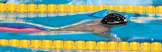 Fotografia de uma pessoa nadando em uma piscina usando touca. O nadador está parcialmente submerso, com a cabeça e um braço aparente fora da água. A piscina tem faixas de boias amarelas flutuando na superfície, separando as raias. A água é azul com ondulações seguindo o movimento do nadador.